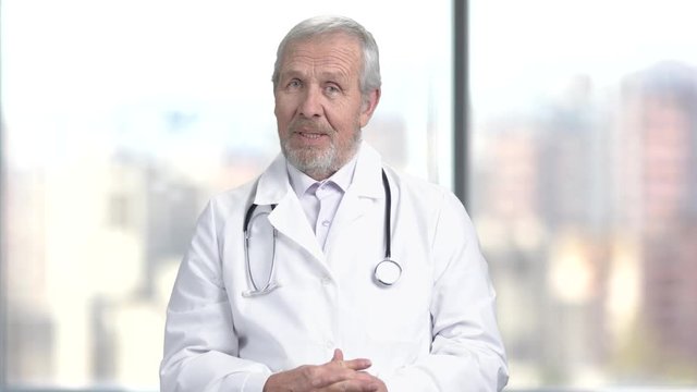 Senior bearded doctor on blurred background. Elderly caucasian doctor is talking and looking at camera. People, professions, medicine.