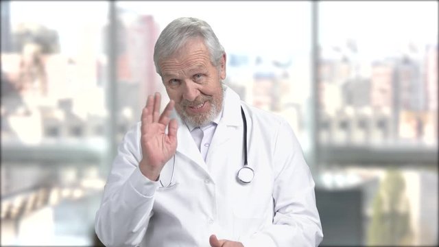 Older doctor talking to camera, portrait. Senior male doctor talking and waving with hand to camera on abstract blurred background.