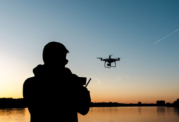 Silhouette of man flying drone by the river at sunset  - obrazy, fototapety, plakaty