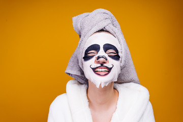a funny young girl with a towel on her head and a white bathrobe smiles, on her face a mask with a...