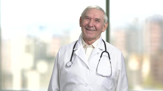 Front view cheerful senior doctor laughing hard. Portrait of a jovial old male doctor in lab coat and stethoscope. Bright abstracrt blurred windows background with view on city.