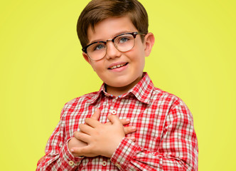 Handsome toddler child with green eyes having charming smile holding hands on heart wanting to show love and sympathy over yellow background