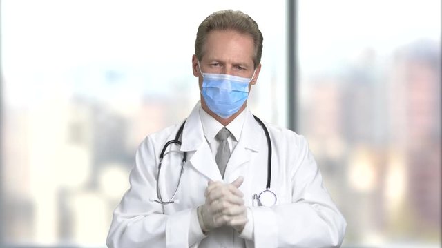 Serious doctor in mask put on gloves. Getting ready to surgery operation. Bright abstract blurred windows background with view on city.