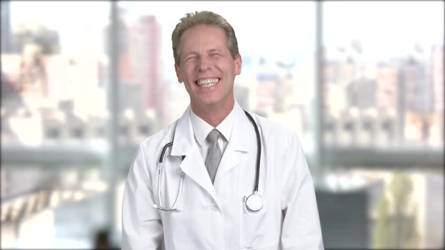 Joyful mature doctor on blurred background. Middle-aged doctor in white coat laughing on window city background.