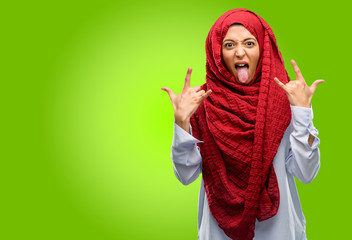 Young arab woman wearing hijab making rock symbol with hands, shouting and celebrating with tongue out