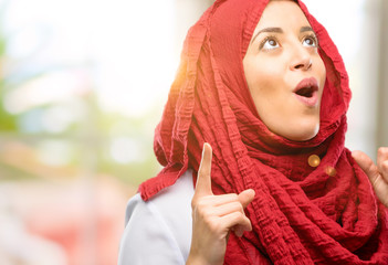 Young arab woman wearing hijab happy and surprised cheering expressing wow gesture pointing up