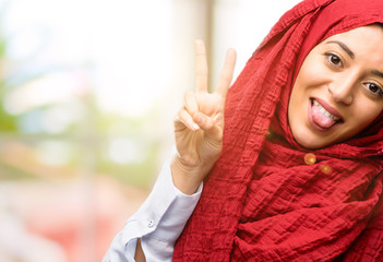 Young arab woman wearing hijab looking at camera showing tong and making victory sign with fingers