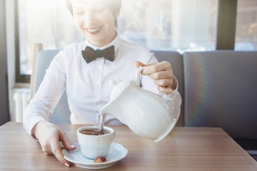 A young pretty girl in a white blouse with a bow tie at a table in a cafe smiles, dreams, pours tea from a teapot into a cup