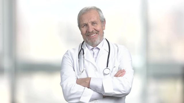 Senior smiling doctor with stethoscope. Portrait of cheerful elderly doctor with crossed arms on blurred background. Happy professional doctor.