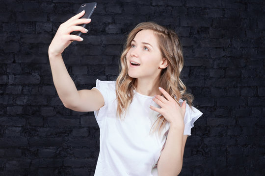 Close up portrait of a young joyful woman holding a smartphone digital camera with hand and making selfie standing against black brick wall background. European beautiful lady takes shot for lover .