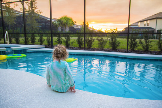 Rear view of a child sitting on the edge of a backyard swimming pool. Concept photo of swimming pool danger and child water safety.