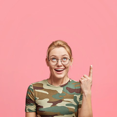 Vertical shot of amazed blonde woman in spectacles, points upwards, sees something great, indicates...