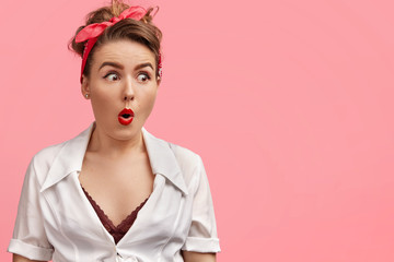 Surprised attractive young female looks with amazed expression, has make up, wears fashionable headband and blouse, isolated over pink background with blank copy space for your advertisment.