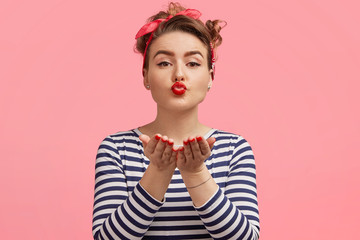 Young beautiful female model blows air kiss, expresses her love to boyfriend, wears striped sweater, isolated over pink background. Horizontal shot of gorgeous woman kisses you with pleasure