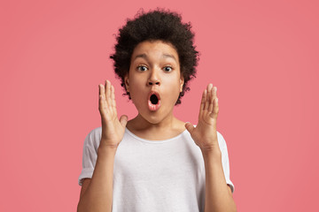 Scared astonished mixed race African male child with bushy hairstyle, gestures with hand as tells...