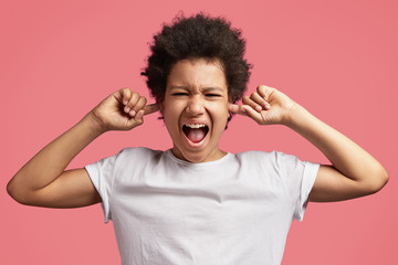 Fototapeta na wymiar Photo of African American mad male child shouts loudly and plugs ears, hears irritated sound, has displeased expression, isolated over pink background. Mixed race teenager with discontent look