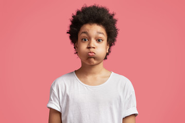 Portrait of small mixed race male kid blows cheeks and puffs breath, has attractive look and bushy Afro hairstyle, keeps silence with friends, wears casual white t shirt, isolated over pink wall - obrazy, fototapety, plakaty