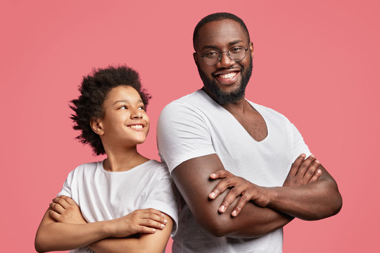 Positive dark skinned male with shining smile keeps hands crossed stands back to attractive African American teenager, have good intention, isolated over pink background. Friendship concept.