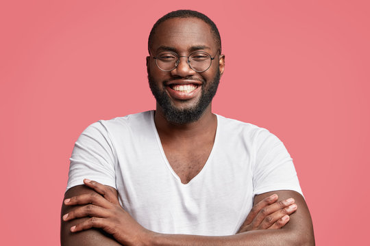 Happy African American male model with dark healthy skin, has broad smile, shows white teeth, keeps hands crossed as demonstrates his confidence, satisfied with result of work, poses indoor.