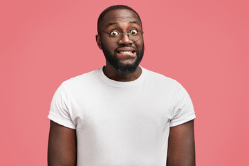 Portrait of surprised African American male in bewilderment, bites lower lip, feels puzzled, wears casual t shirt and spectacles, isolated over pink background. Dark skinned young man poses indoor