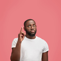 Vertical shot of dark skinned serious confident African male points upwards with fore finger,...