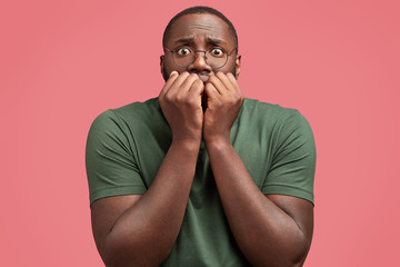 Candid shot of handsome African American male looks with worried expression, bites nails, feels anxious and nervous as being scared of something or sees phobia, poses against pink background