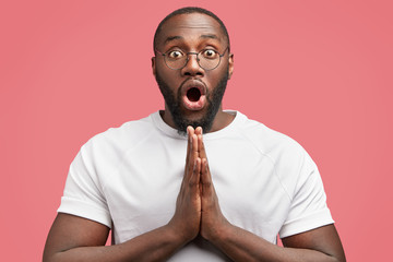 Portrait of emotional surprised young African American guy with shocked expression, keeps palms pressed together, feels amazed, has plump figure, being unshaven, isolated over pink background