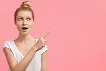 Horizontal portrait of young beautiful blonde female model with European appearance, looks in bewilderment away, indicates at blank copy space for your advertising content or promotional text