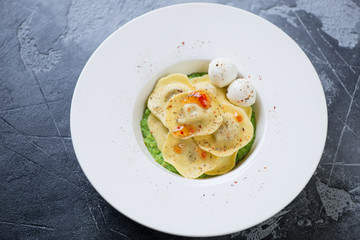 White plate with ravioli on a bed of green peas puree, mozzarella and sauce. Elevated view on a grey stone background, studio shot