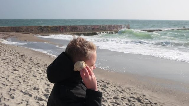 Cute funny little kid enjoying beach holidays outside on cold windy spring day. Child dreaming about warm summer standing at sandy beach and listening seashell.