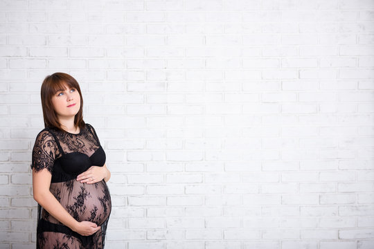 pregnant woman in black lace dress thinking or dreaming about something over white brick wall background