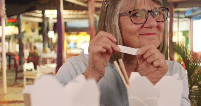 Happy elderly woman showing off her fortune cookie confidently sitting at outdoor table, Portrait of happy senior woman reading fortune after eating Chinese food outside, 4k