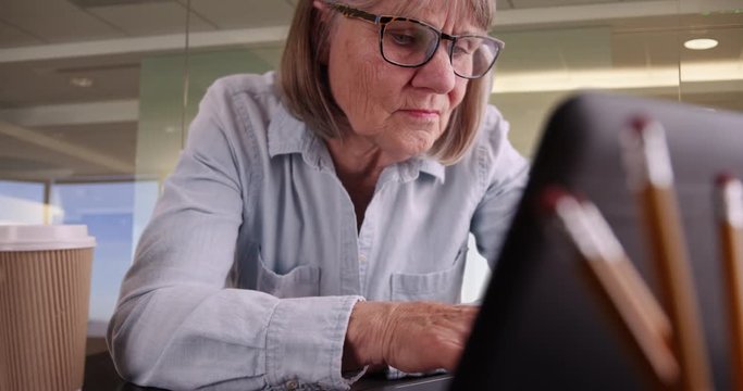 Close-up of senior woman typing on laptop working in corporate workplace, Portrait of older woman using portable computer in modern office space setting, 4k