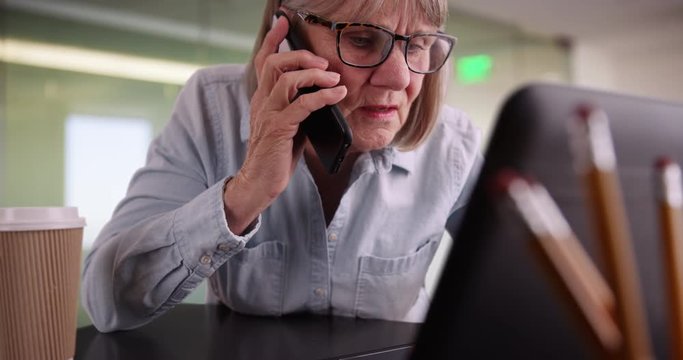 Close-up portrait of elderly woman talking on cellphone in modern office setting, Closeup of senior woman employee on phone with client and working on laptop, 4k 