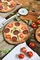 Mini Pizza With Cherry Tomatoes And Salami Sausages On A Wooden Background Natural Rustic, A Pizza Cutter And Ingridienty.