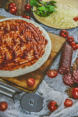 Ingredients for Pizza: basis dough, salami sausage, cherry tomatoes, grated cheese, tomato paste, herbs and arugula. Natural Wooden Background With Linen Napkin and Knife For Pizza