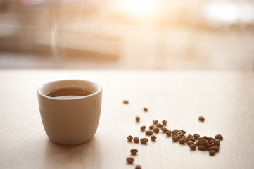 The window in which the sun is shining, on the table, a hot Cup of coffee from which steam rises and near are roasted coffee beans. Side view with copy space for your text