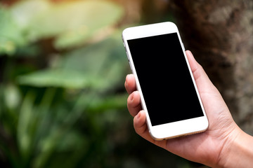 Mockup image of a hand holding white smart phone with blank black desktop screen in outdoor with blur green nature background