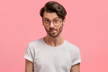Fototapeta premium Horizontal portrait of serious unshaven male manager looks with thoughtful hesitant expression, curves lips, tries to find solution in mind, wears casual white t shirt, isolated over pink background