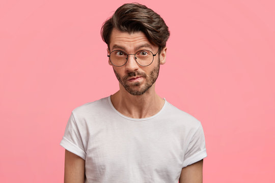 Horizontal portrait of serious unshaven male manager looks with thoughtful hesitant expression, curves lips, tries to find solution in mind, wears casual white t shirt, isolated over pink background