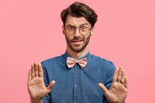 Elegant male youngster dressed in denim shirt with pink bowtie, gestures with discontent expression in studio, demonstrates his refusal and disagreement. Stylish male shows stop sign indoor, says no