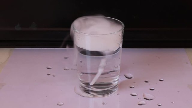 Dry ice in a glass of water, it moves and engender air bubble with white smoke on white floor with black background.