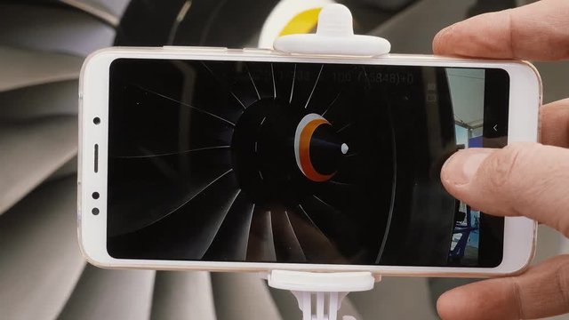 A student of a technical institute is studying an aircraft engine. Point of view. A man's hand takes a snapshot of a jet engine using a modern smartphone and selfie monopod.