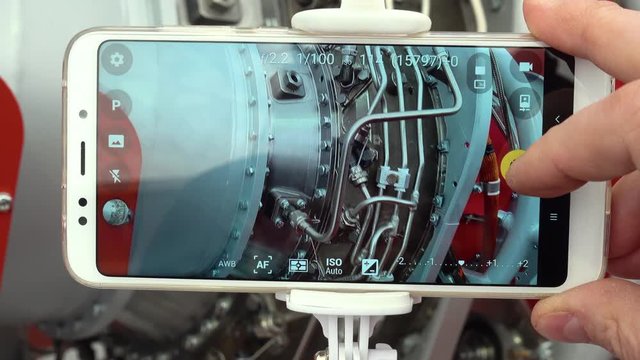 A student of a technical institute is studying an aircraft engine. Point of view. A man's hand takes a snapshot of a jet engine using a modern smartphone and selfie monopod.