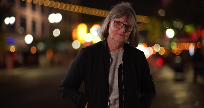 Somber old lady with hands in pockets looking at camera standing in city street, Serious caucasian woman with hair blowing in wind posing in street with bokeh lights in background, 4k