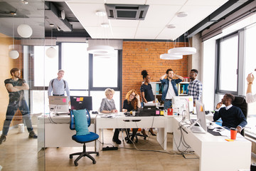 young Caucasian bearded guy in white T-shirt and denim jacket has come back from vocation and talking about his pastime to co-workers while two attractive women sitting and ignoring him.