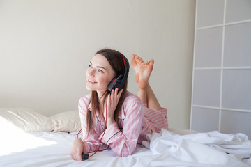 brunette in pink Pajamas listening to music with headphones on the bed