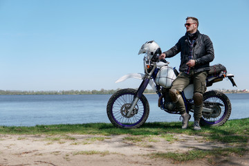 Obraz premium Young stylish man sit on classic retro off road track motorcycle on the beach, outdoor portrait, posing, in lather jacket and Sunglasses, travel active lifestyle concept, ocean, sea, lake, river