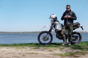 Fototapeta na wymiar Young stylish man sit on classic retro off road track motorcycle on the beach, outdoor portrait, posing, in lather jacket and Sunglasses, travel active lifestyle concept, ocean, sea, lake, river
