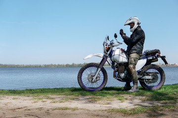 Plakat Young stylish man sit on classic retro off road track motorcycle on the beach, outdoor portrait, posing, in lather jacket and Sunglasses, travel active lifestyle concept, ocean, sea, lake, river